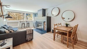 Uma sala de estar moderna com lareira, uma área de jantar com cadeiras de madeira e grandes janelas com vista para árvores.