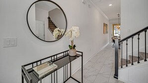 A hallway with a round mirror, a console table with a vase of white flowers, a staircase with black railings, and a dining area in the background. - EV_4228 - 4BR / 3BA Storey Lake Townhome near to Disney, Private Pool & Resort (Kissimmee)