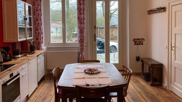 A kitchen with a dining table, wooden cabinets, and a window with curtains. - No letting via FEWO (Lärz)