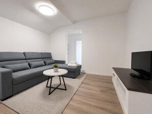 Un salon moderne avec un canapé gris, une petite table ronde ornée d’une plante, un téléviseur à écran plat et un plancher en bois.