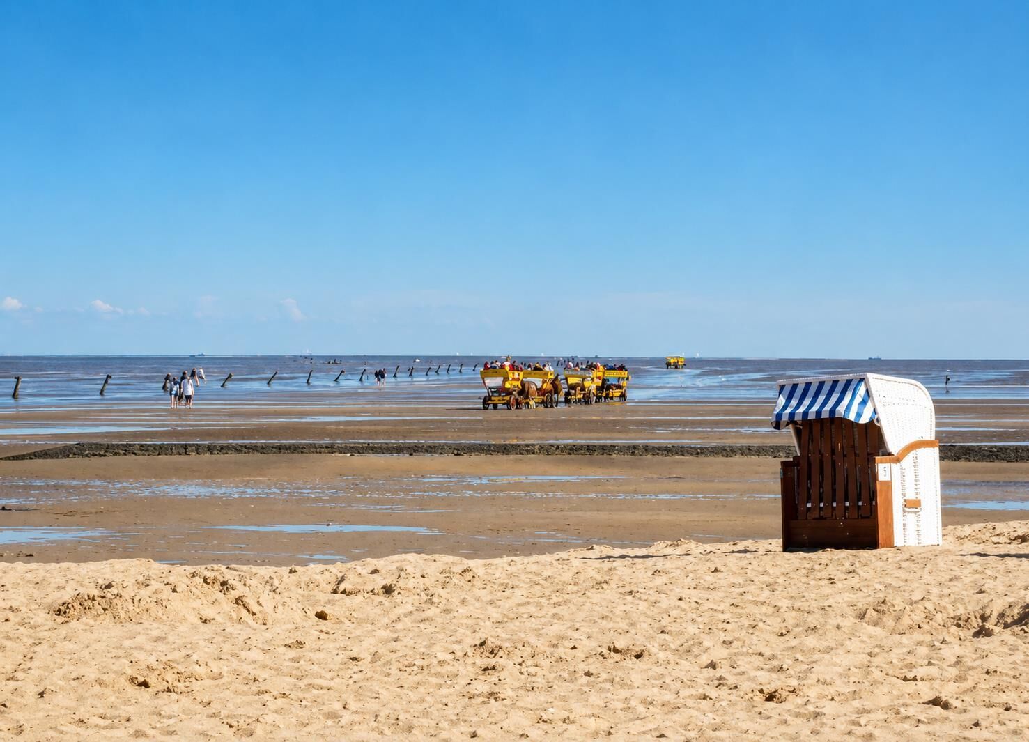 A beach with a sandy foreground, a wooden structure with a striped awning, and a group of yellow horse-drawn carriages in the background.