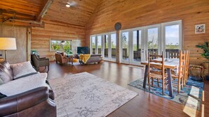A spacious living room with a wooden ceiling, a large window, a sofa, a coffee table, and a dining area with chairs and a table. - Beautiful log cabin w/ private dock, gameroom, 23mi to beach, 3bd2ba, sleeps 11 (Youngstown)