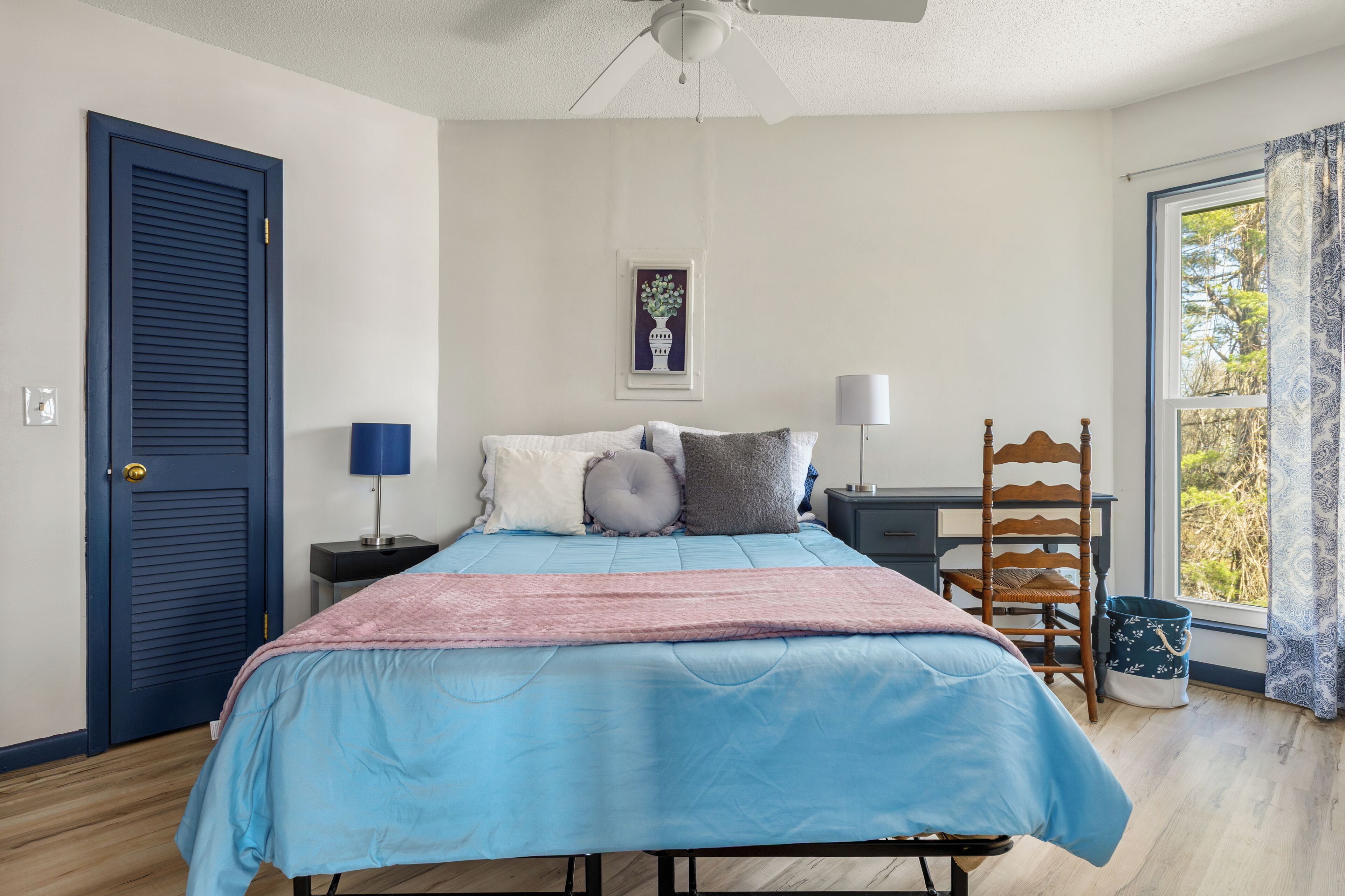 A bedroom with a bed, a desk, a chair, a window with a view, and a door.