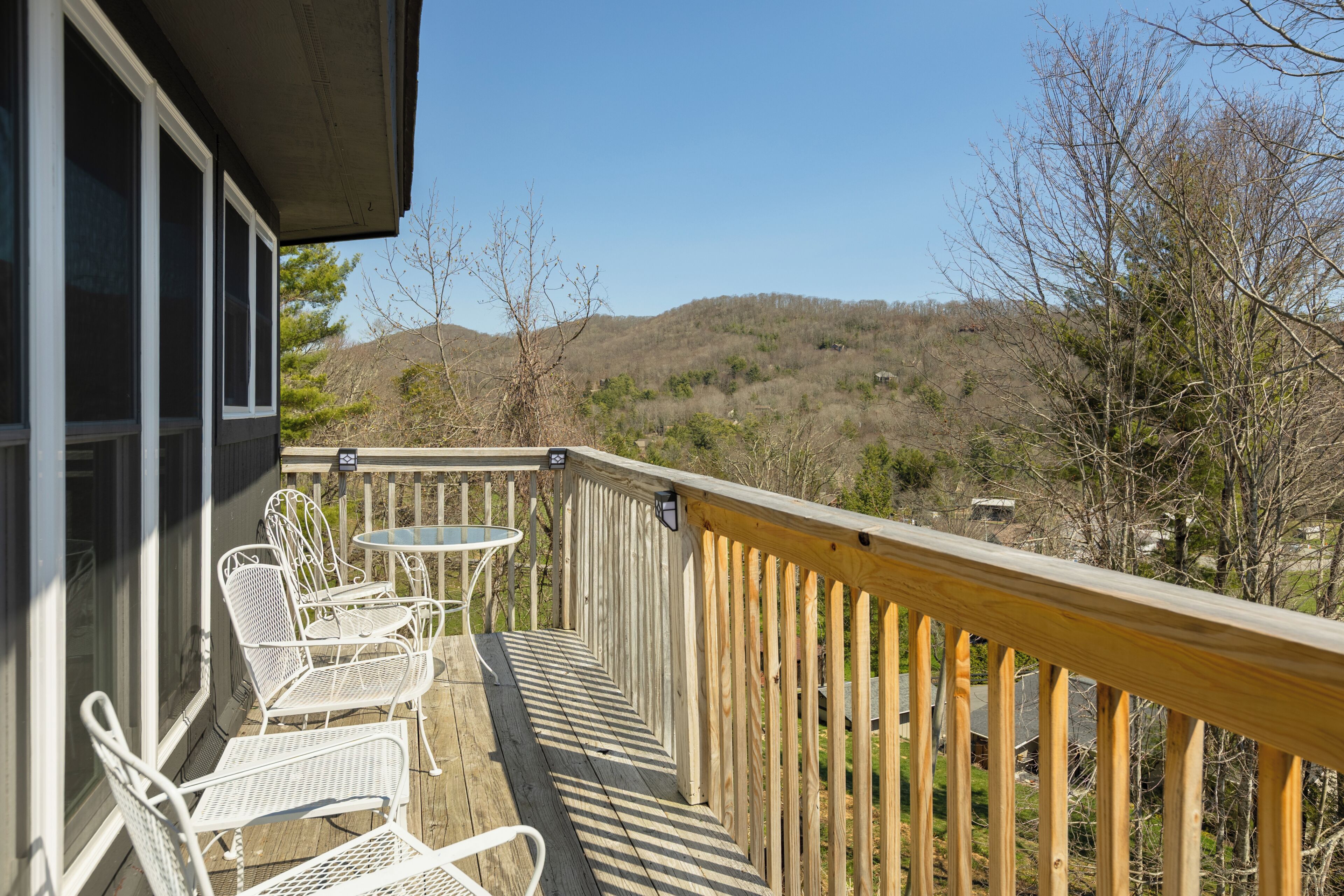 A wooden deck with white metal chairs and a small table, overlooking a hilly landscape.