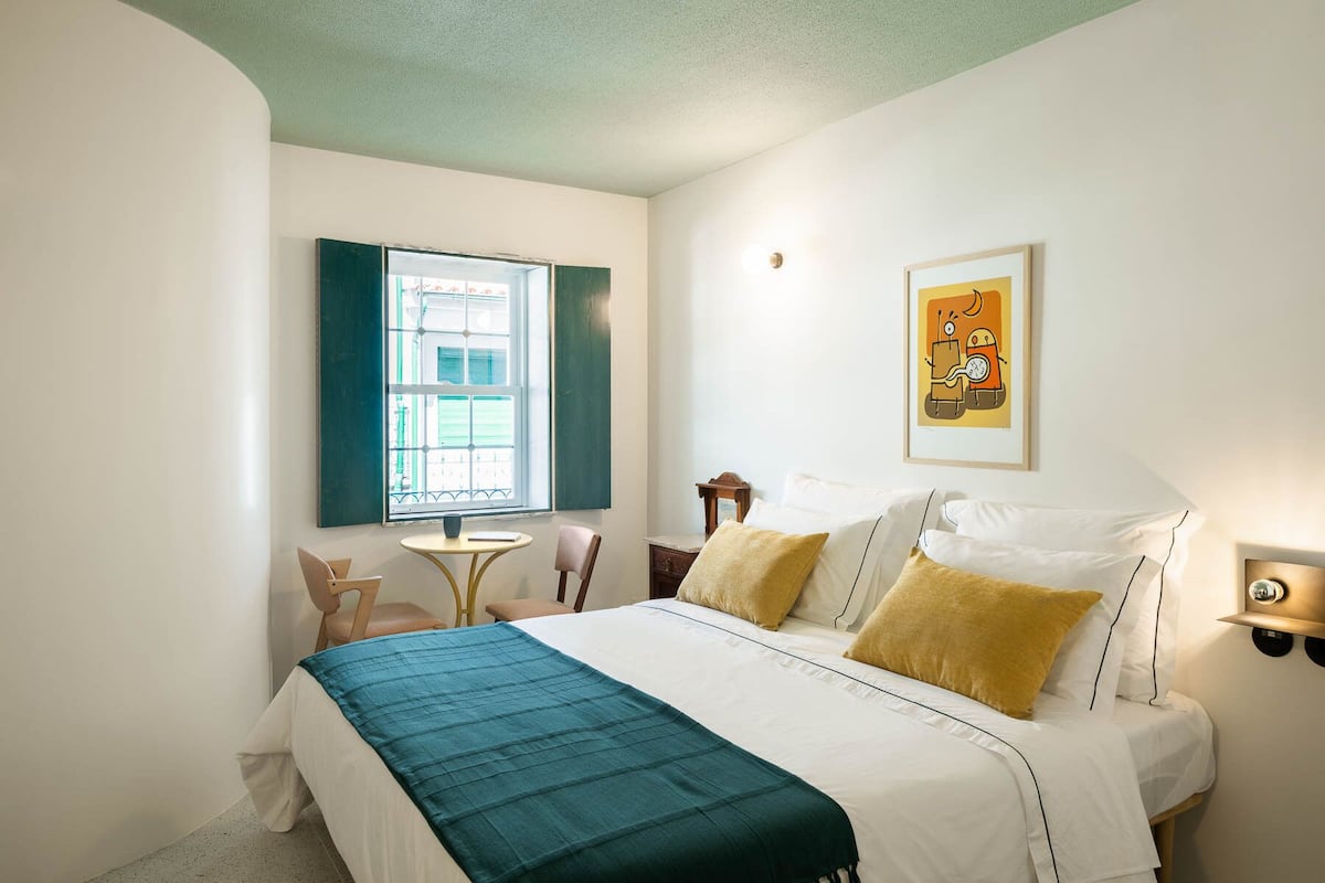 Un dormitorio con cama, una mesita, dos sillas, una ventana y una lámpara fijada a la pared.