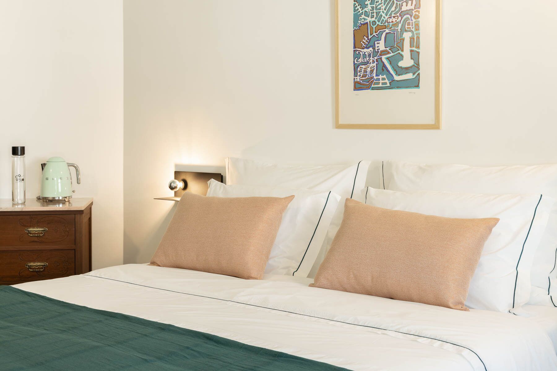 Cama bem arrumada com duas almofadas, mesa de cabeceira com chaleira e luminária na parede.