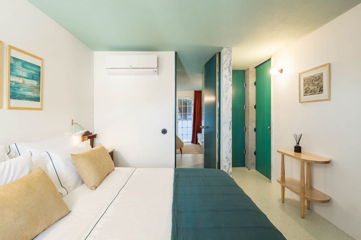 Un dormitorio con cama, mesita de noche, aire acondicionado de pared y puerta que da a otra habitación.