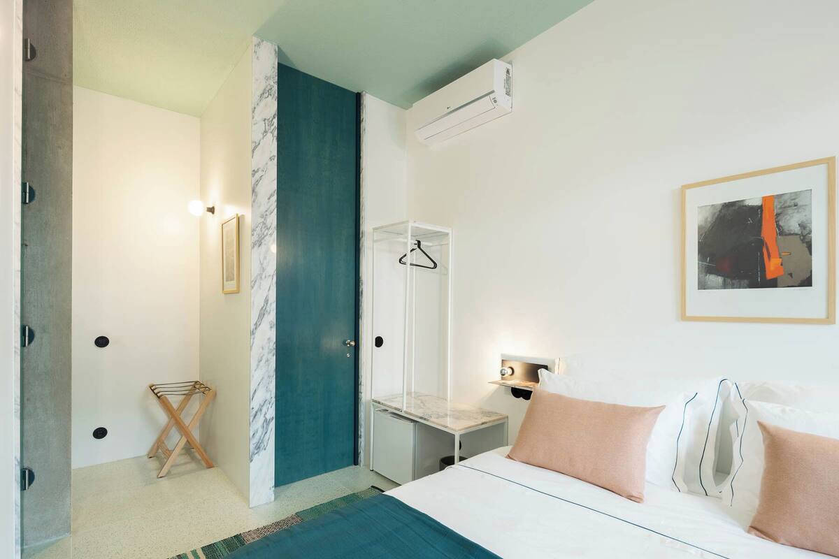 Una habitación de hotel con cama, mesita de noche, lámpara colgante, una obra de arte enmarcada y zona de ducha.