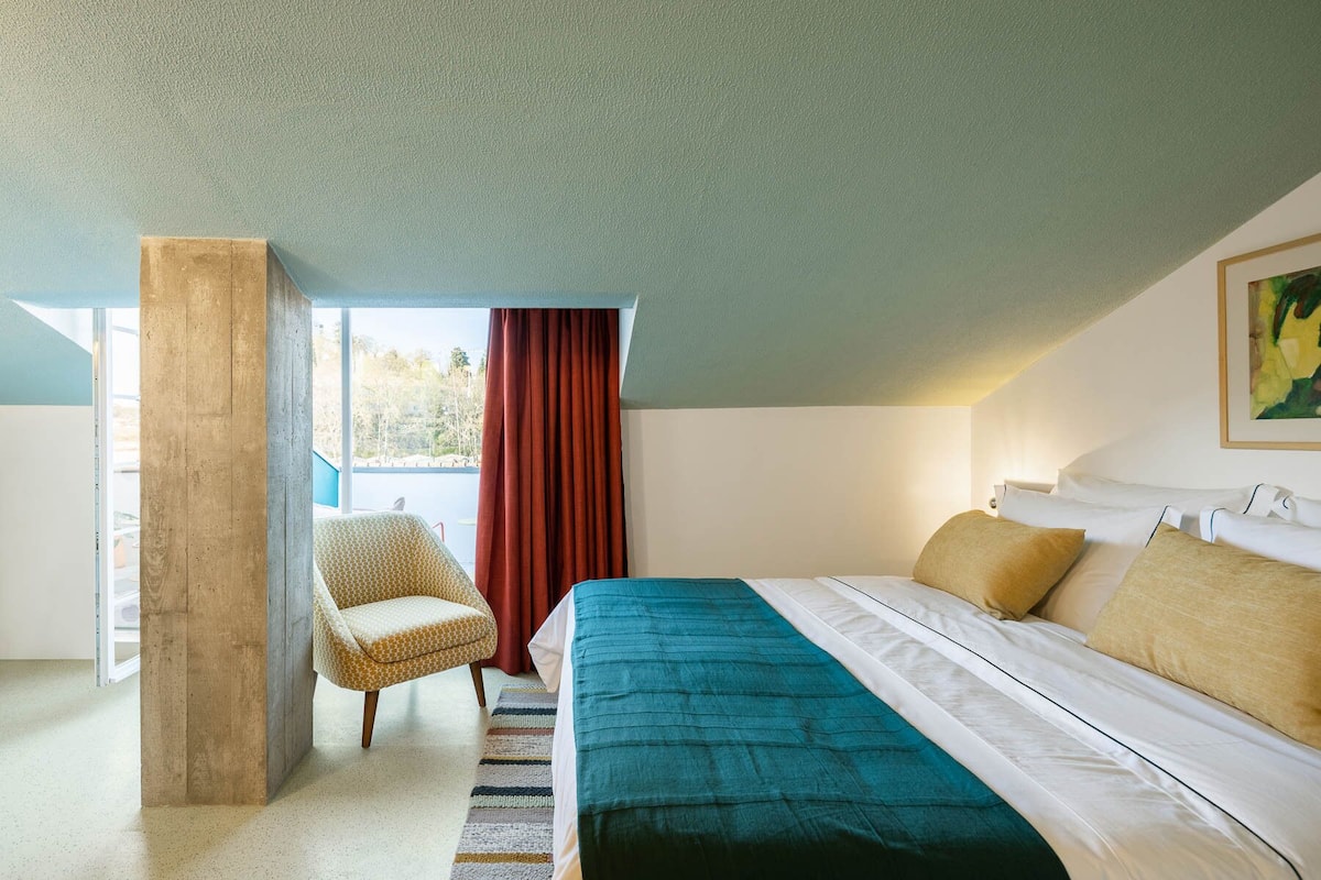 Un dormitorio con cama, una silla, ventana con cortinas y un cuadro en la pared.