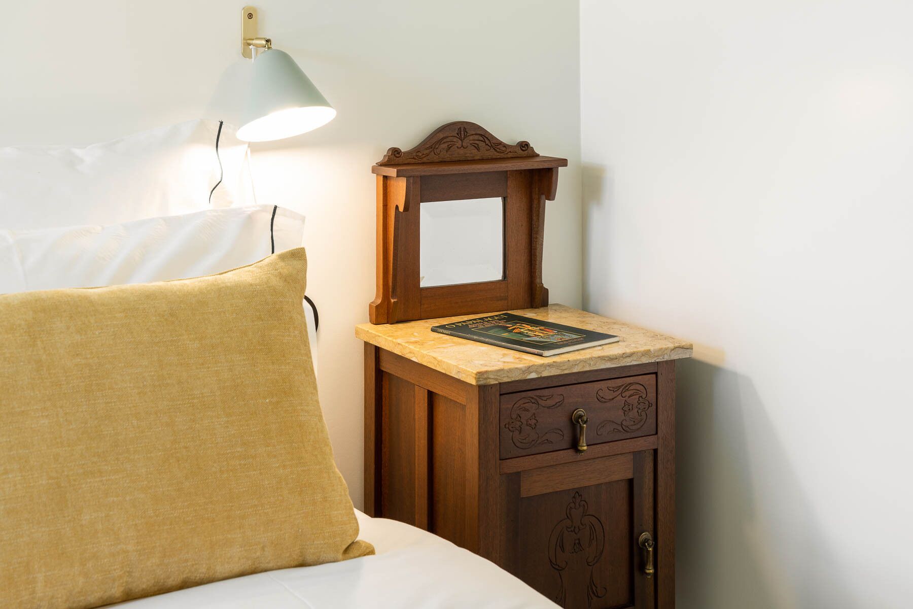 A neatly made bed with a beige pillow, a small wooden nightstand with a lamp, and a book on top.