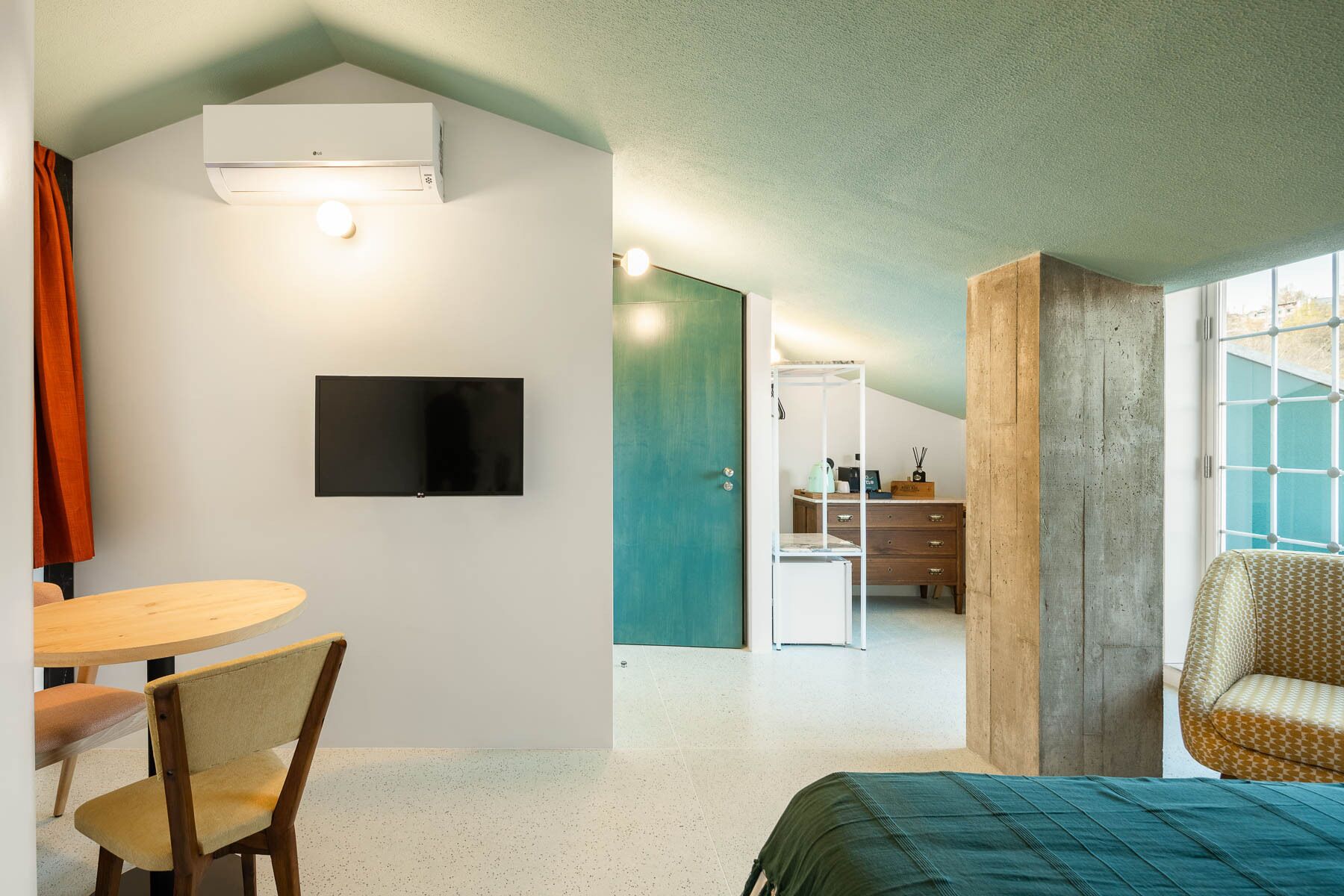 A modern hotel room with a flat-screen TV, a dining area with a round table and chairs, a bed with a green bedspread, and a large window.