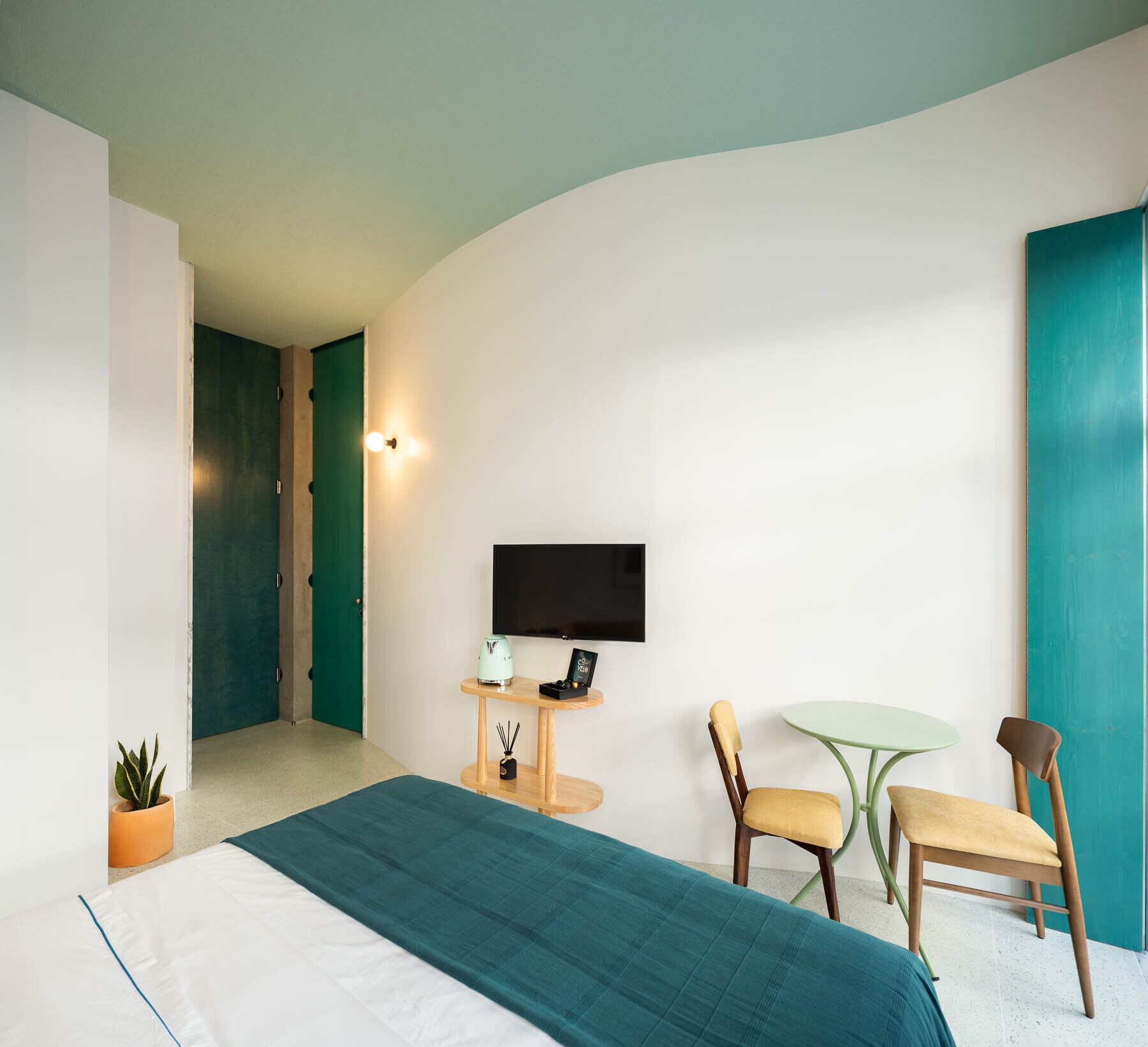 Una habitación de hotel moderna con una cama, un escritorio con televisor de pantalla plana, dos sillas, una mesita y una planta en maceta.
