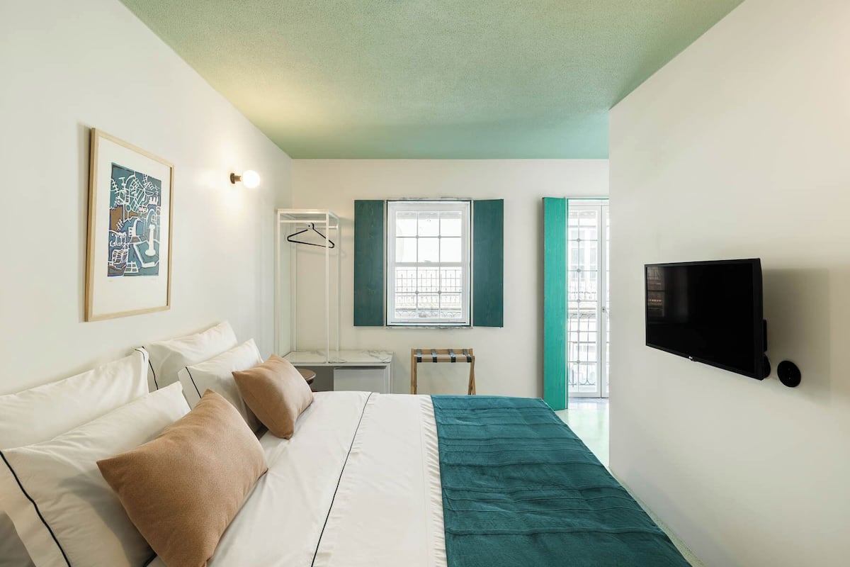 Un dormitorio con cama, televisión, ventana con contraventanas y lámpara colgante.