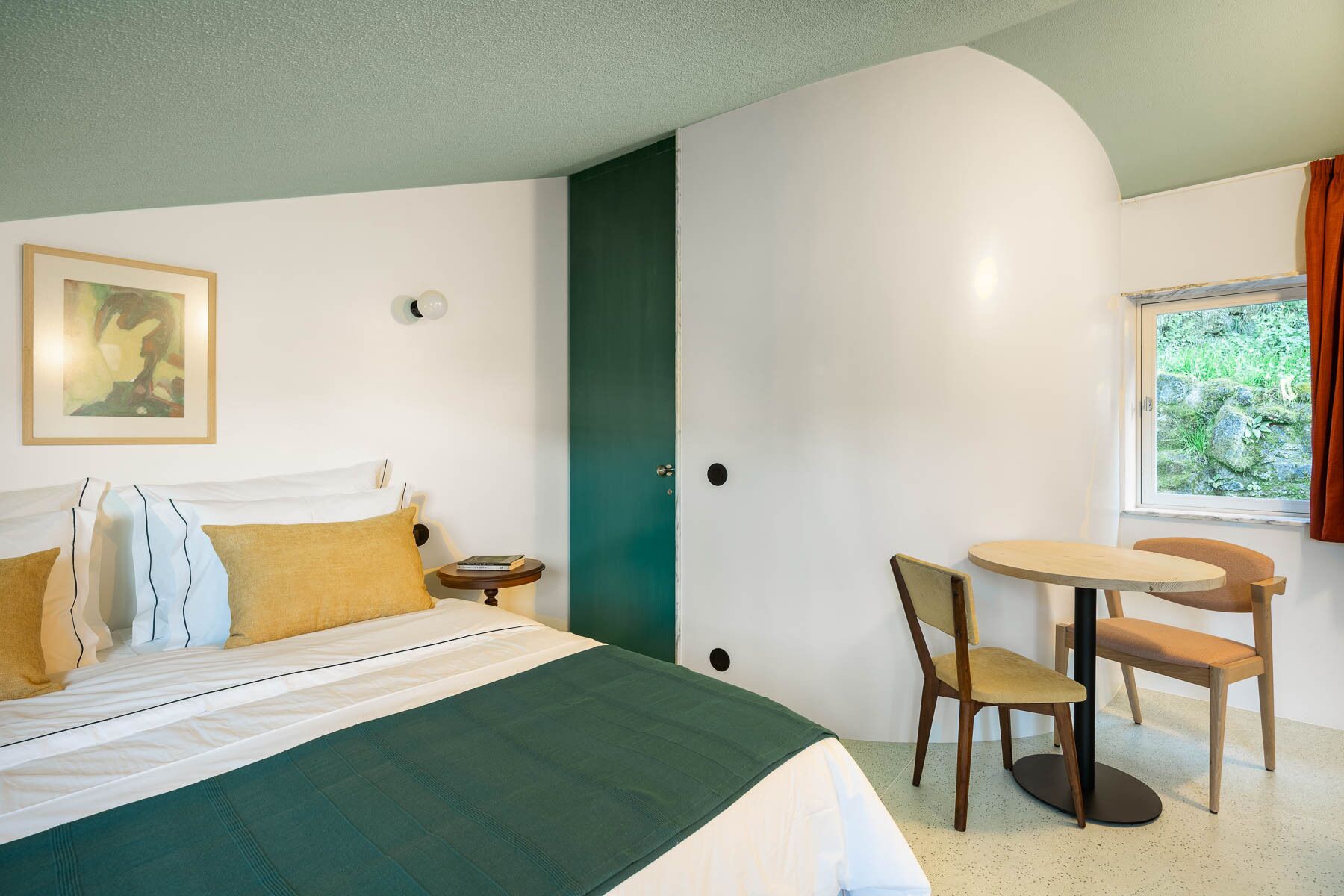 Une chambre à coucher comprenant un lit, une table de chevet, une chaise, une table ronde et une fenêtre donnant sur un paysage verdoyant.