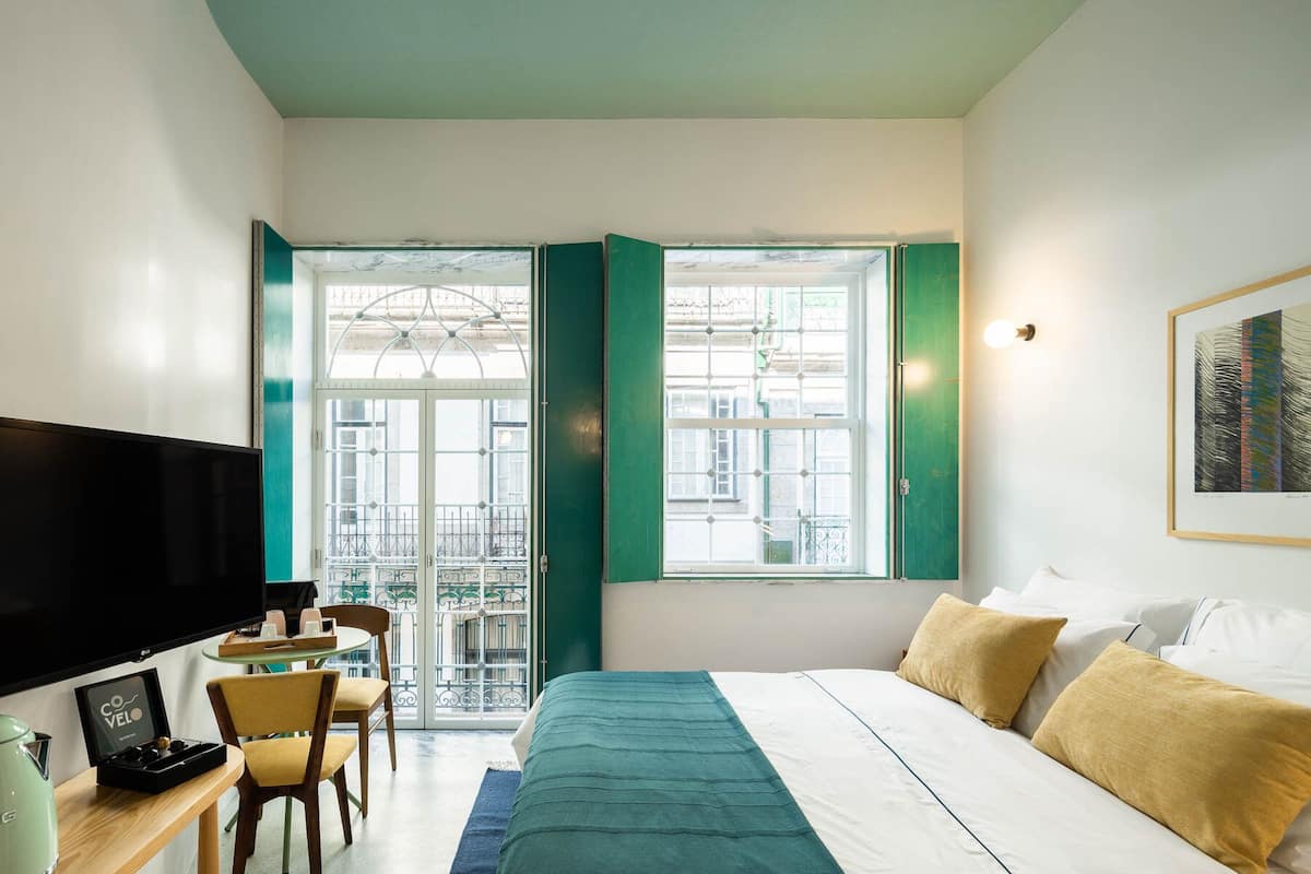 Habitación de hotel con cama, televisor, escritorio con silla y ventana con vistas a edificios.