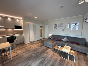 Uma sala de estar moderna com um sofá cinzento, uma pequena mesa de centro e uma área de jantar com uma mesa e cadeiras brancas.