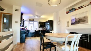 A modern kitchen with a dining area, featuring a wooden table and chairs, a built-in oven, and a hanging light fixture. - Stones throw from the sea (Seaford)