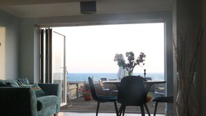 Ett öppet vardagsrum med utsikt över havet, ett matbord med stolar och en soffa med kuddar.