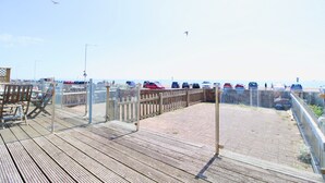 Ett trädäck med ett räcke som vetter ut mot ett strandområde med parkerade bilar och en klar himmel.