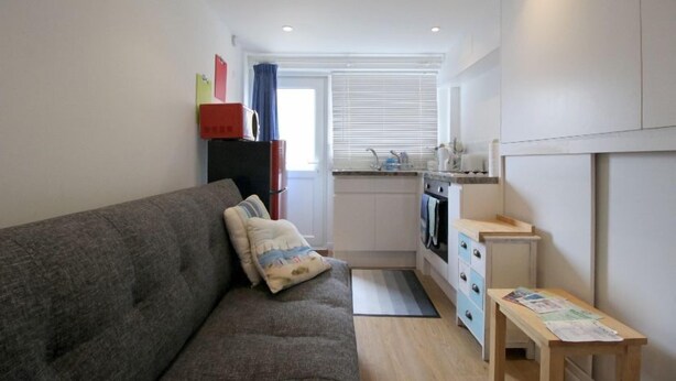 Un espacio habitable compacto con sofá, una mesita, kitchenette y ventana con persianas.