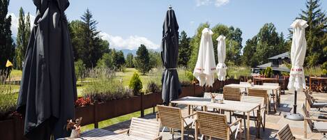 An outdoor patio with wooden tables and chairs, covered by umbrellas, overlooking a landscaped garden and distant mountains.