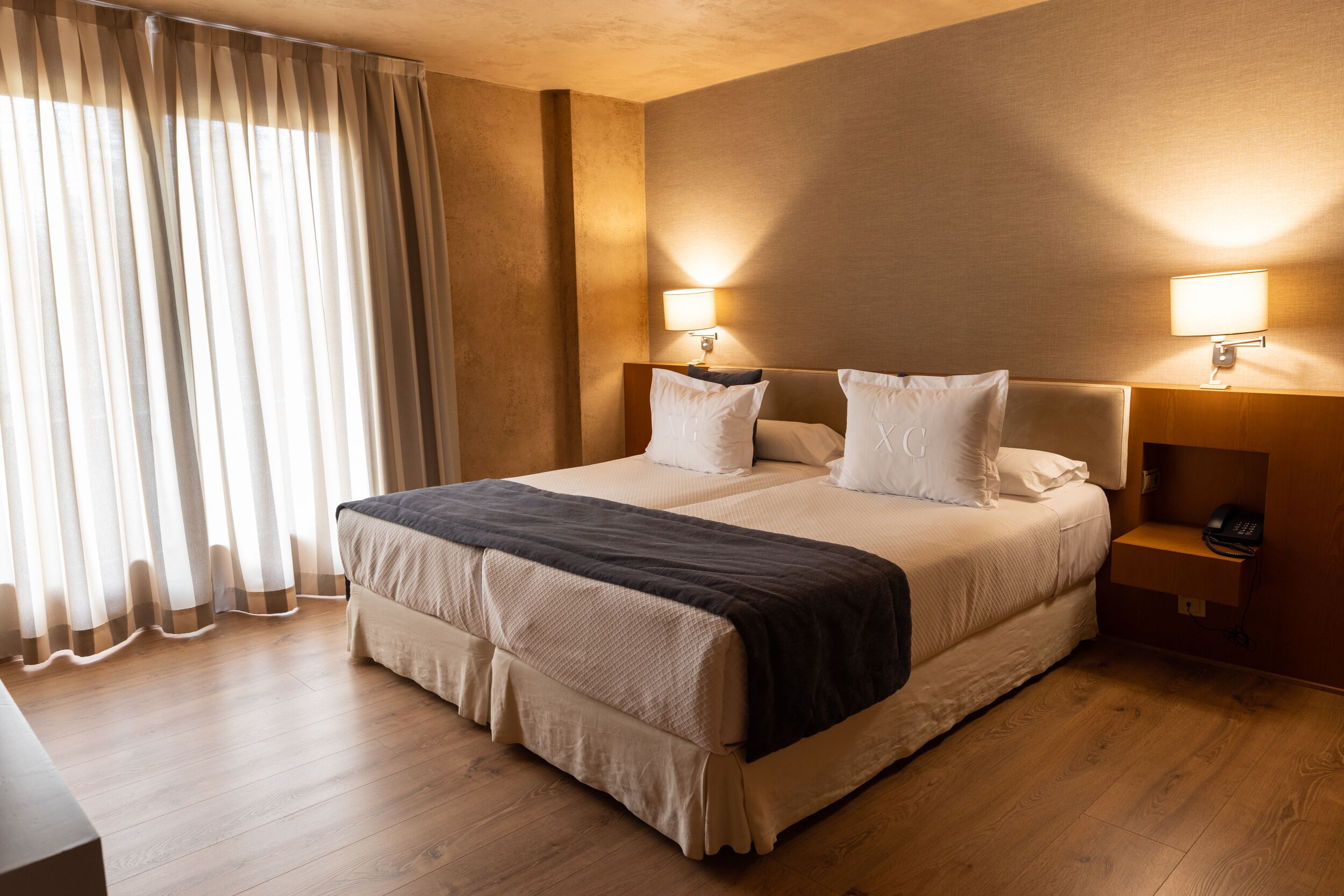 A hotel room with a large bed, two bedside lamps, a wooden nightstand, and a window with sheer curtains.