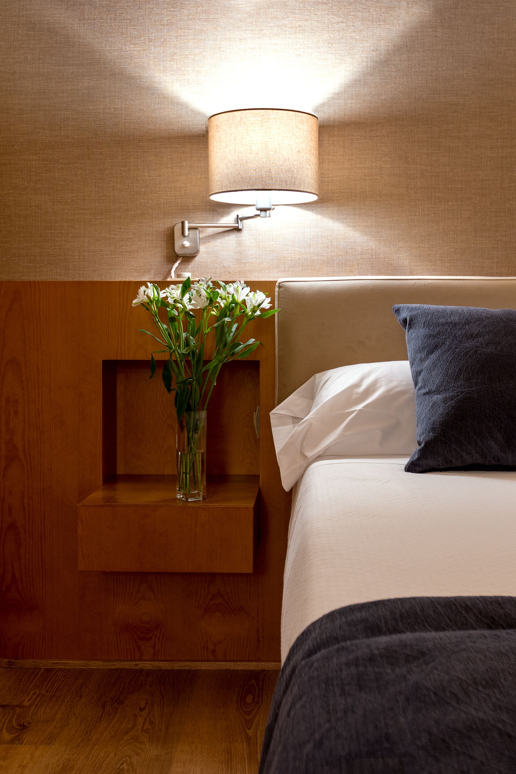 Een netjes opgemaakt bed met een donkeroog en een wit kussen, een nachtkastje met een vaas witte bloemen, en een wandlamp.