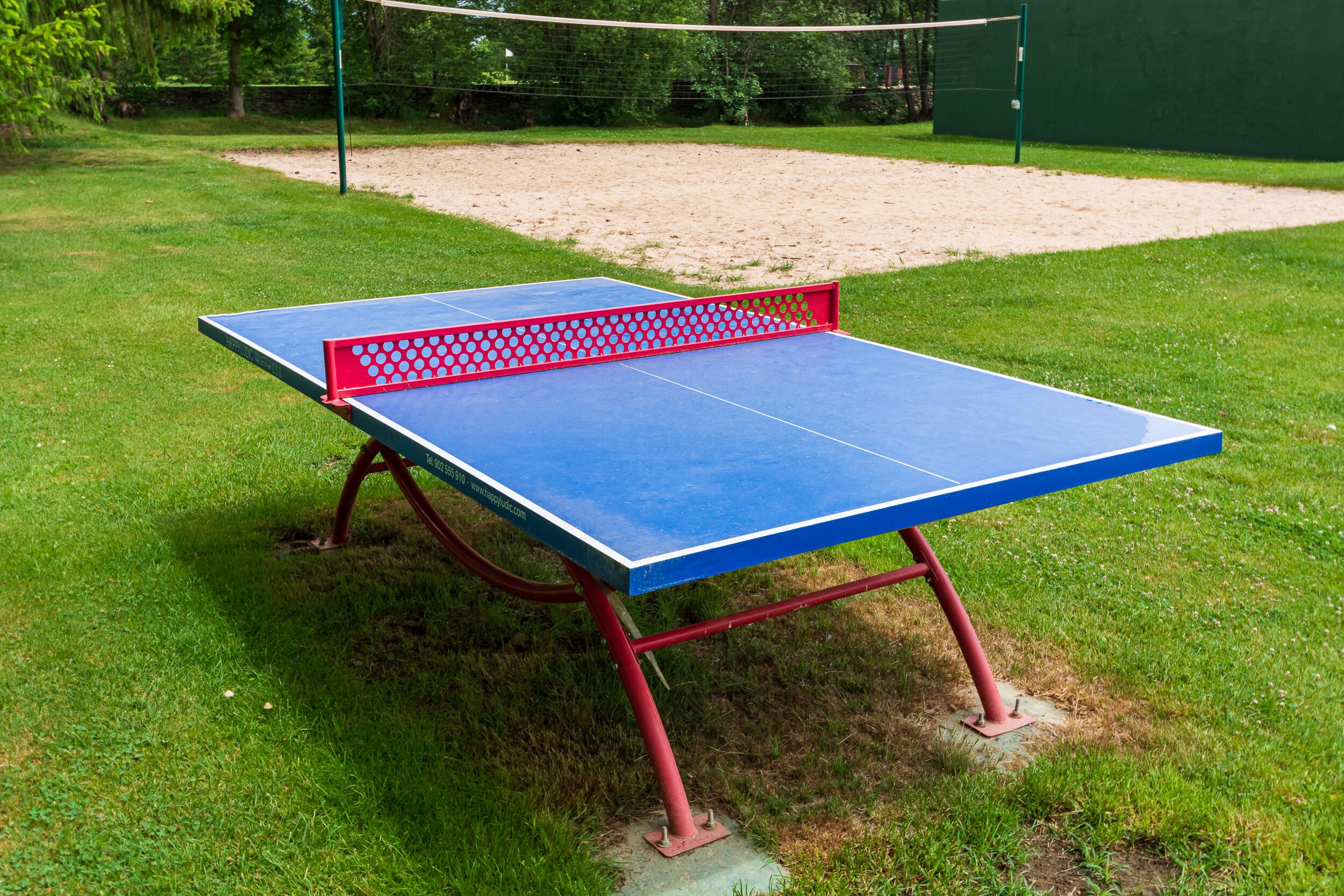 Una mesa de ping pong con patas rojas y superficie azul, ubicada en un césped con un arenero al fondo.