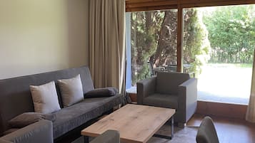 A modern living room with a grey sofa, a wooden coffee table, and two armchairs. Large windows provide a view of trees and a patio.