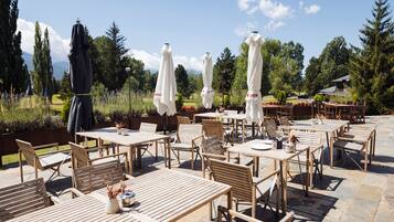 Un patio al aire libre con mesas y sillas de madera, sombrillas blancas y vistas a árboles y un paisaje distante.