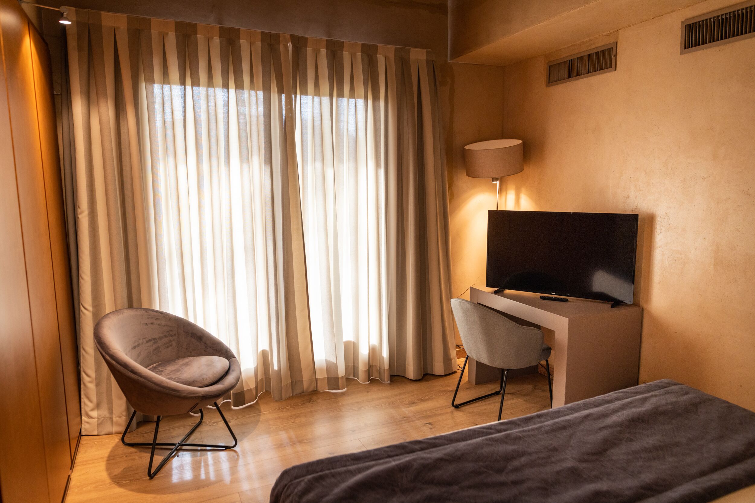 Ein Hotelzimmer mit einem Bett, einem Schreibtisch mit Fernseher, einem Sessel und großen Fenstern mit Vorhängen.