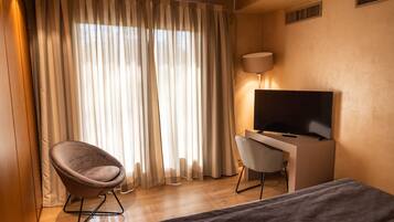 Ein Hotelzimmer mit einem Bett, einem Schreibtisch mit Fernseher, einem Sessel und großen Fenstern mit Vorhängen.