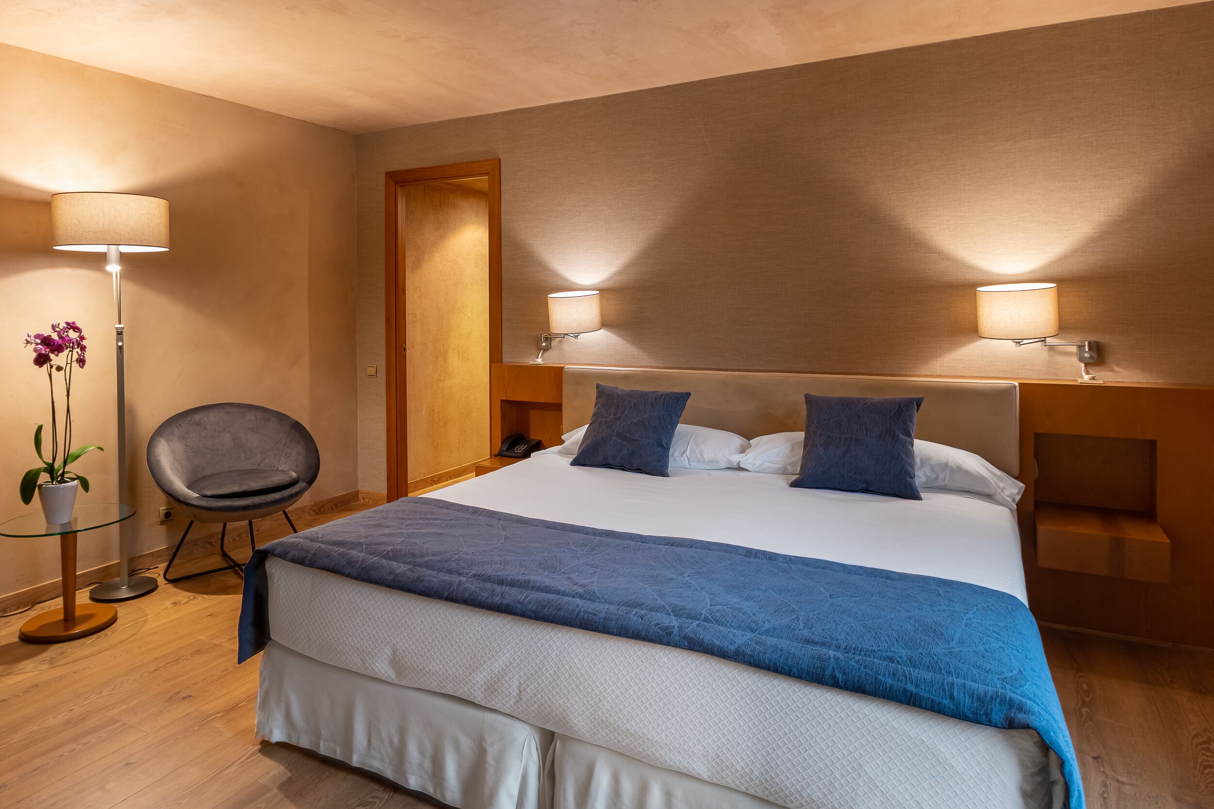 Un cuarto de hotel ordenado con una cama grande, dos lámparas de noche, una silla y una mesita con una planta.