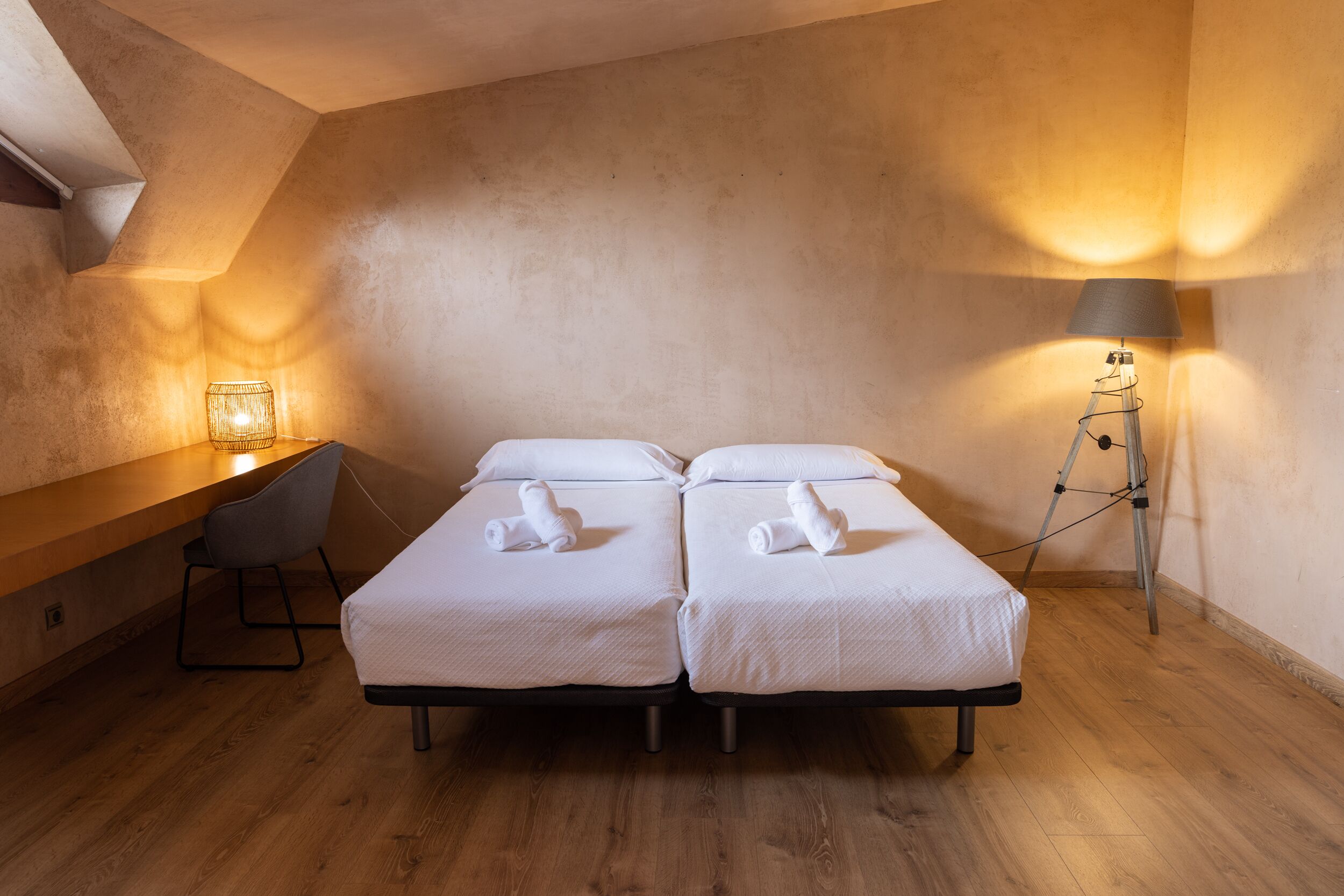 A room with two single beds, a desk, a chair, a lamp, and a wooden floor.