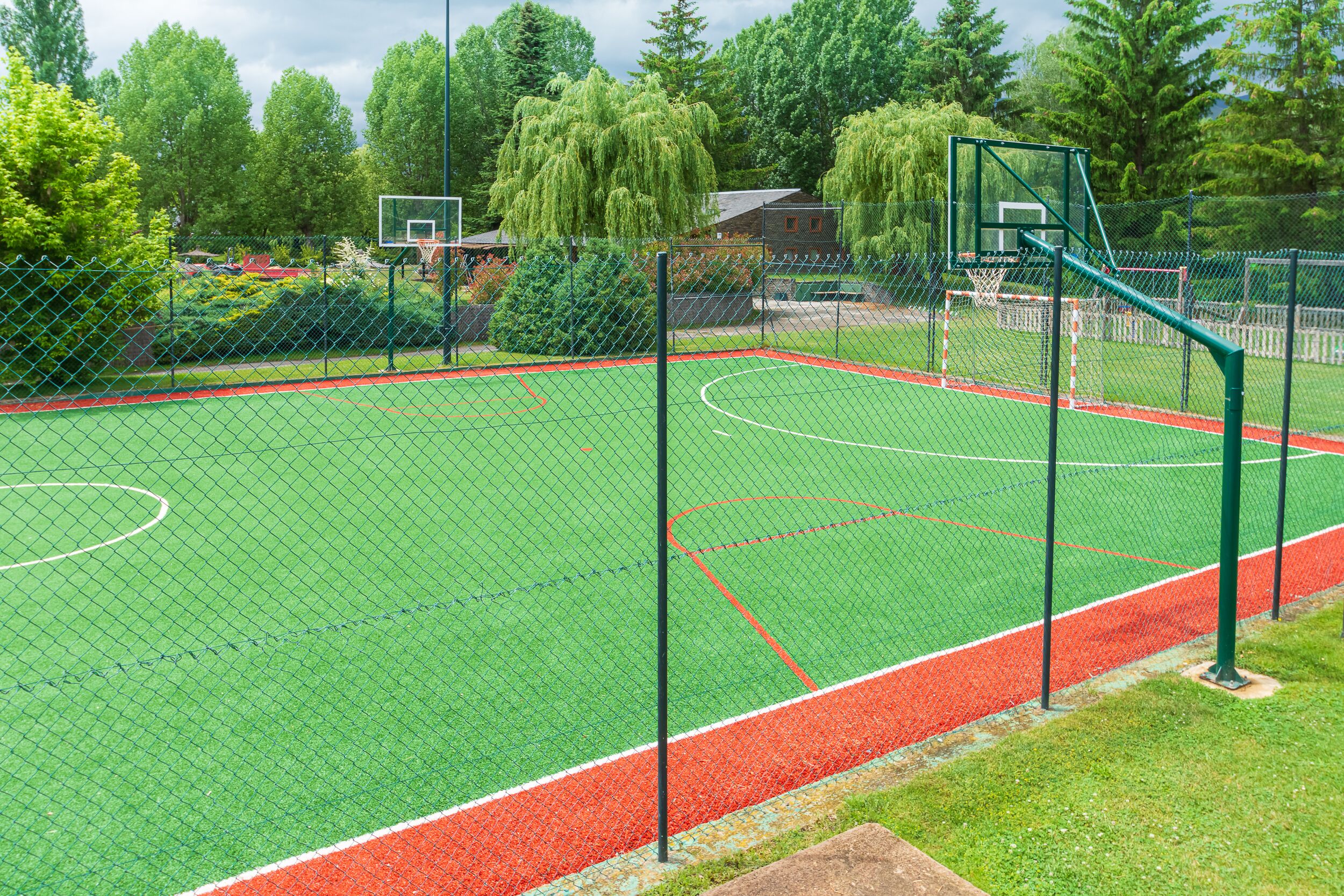 A basketball court with a green surface, surrounded by a chain-link fence, and trees in the background.