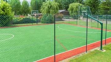A basketball court with a green surface, surrounded by a chain-link fence, and trees in the background.