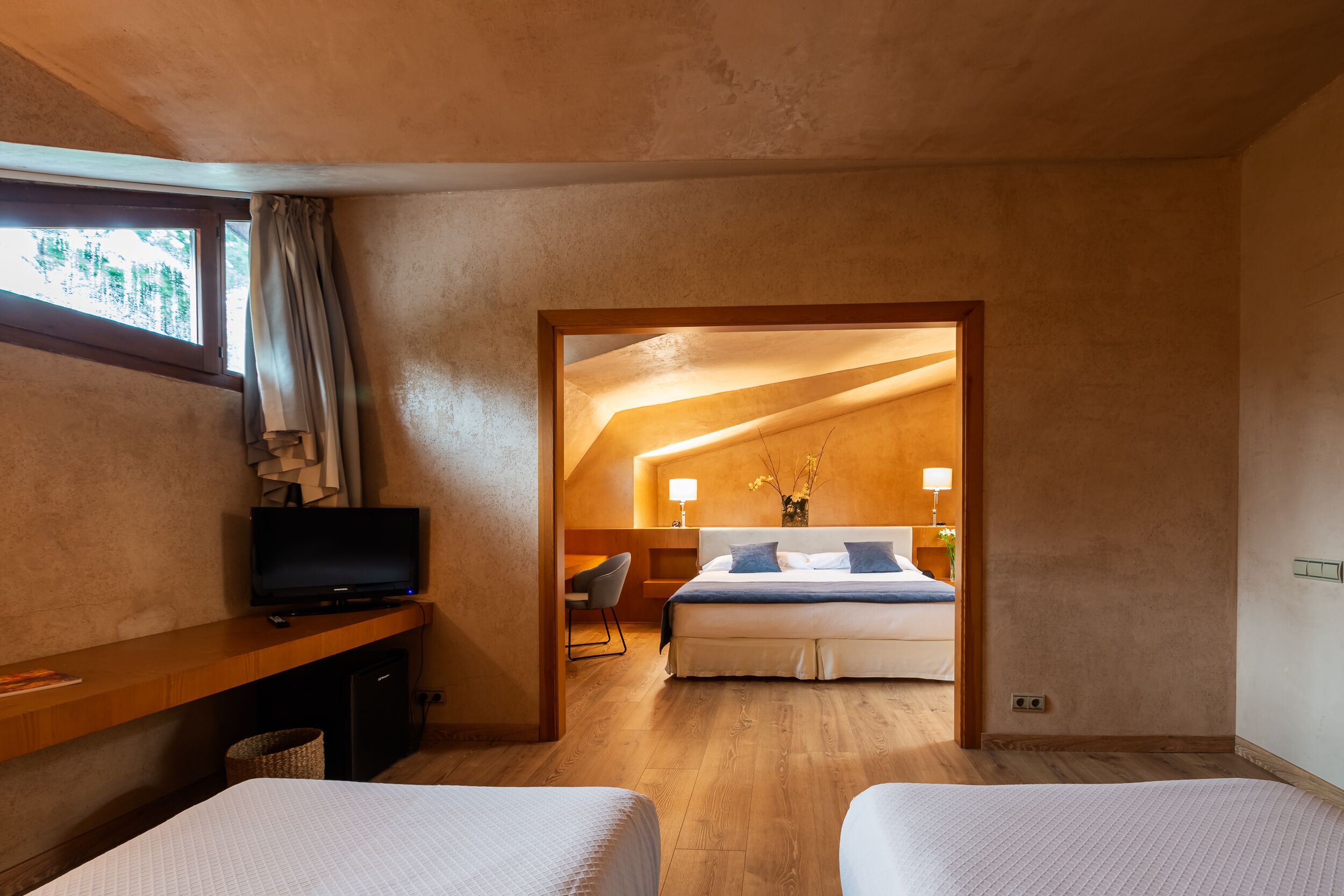 A hotel room with a wooden desk, a television, a bed with a blue bedspread, and a view of another room through an open doorway.