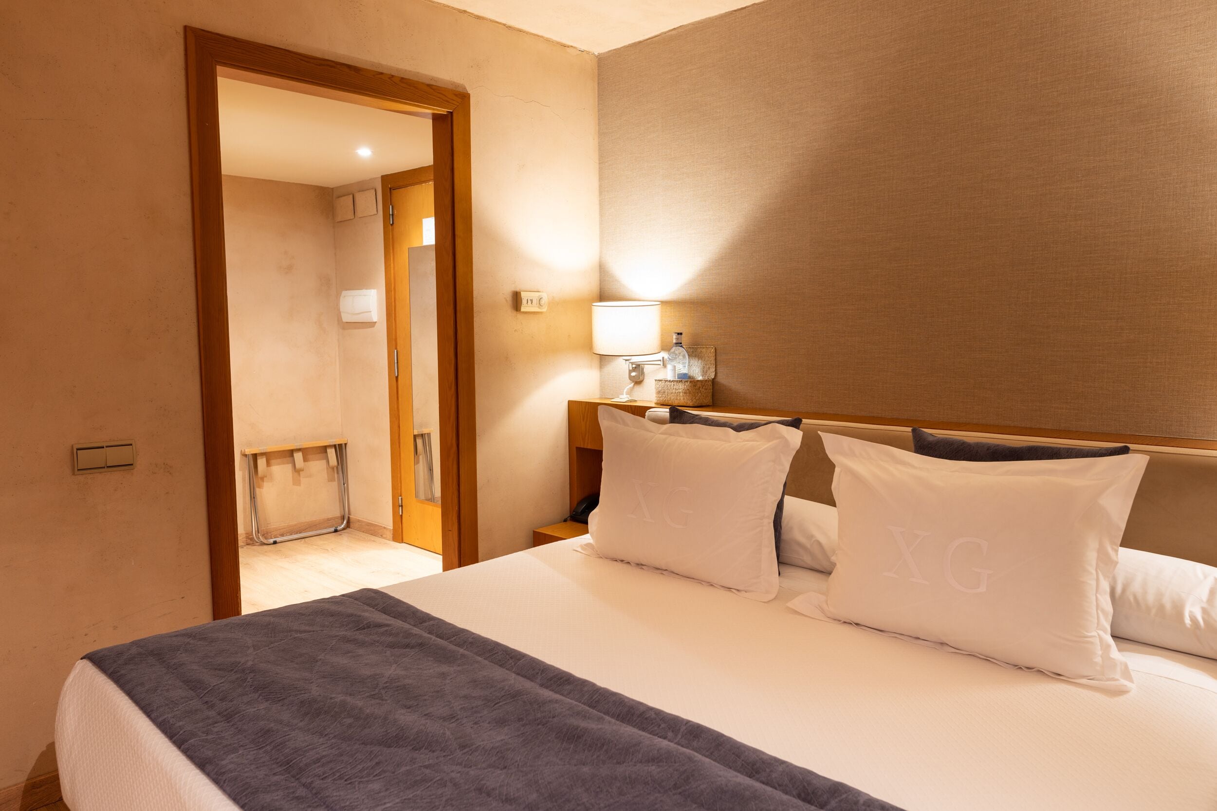 Una habitación de hotel con cama, mesita de noche, lámpara y puerta que da a un baño.