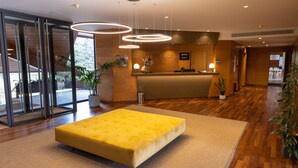 A reception area with a yellow tufted sofa, a wooden reception desk, and a large potted plant. - Hotel Xalet del Golf (Bolvir)