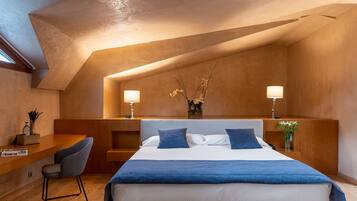 Un dormitorio moderno con techo inclinado, una cama grande con ropa de cama azul, dos lámparas de noche y un escritorio pequeño con una silla.