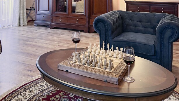 A room with a large wooden cabinet, a blue armchair, a round coffee table with a chessboard and wine glass, and a chandelier.