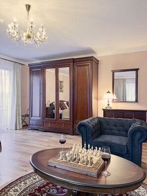 Une pièce comprenant un grand meuble en bois, un fauteuil bleu, une table basse ronde avec un jeu d’échecs et un verre à vin, et un lustre.