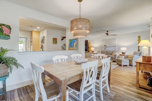 Un coin repas avec une table en bois et des chaises blanches, un luminaire suspendu en osier, et une vue sur le salon, où l’on aperçoit un canapé et un ventilateur de plafond.