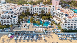 Un complejo turístico con varias edificaciones, una piscina central y numerosas tumbonas en la playa.