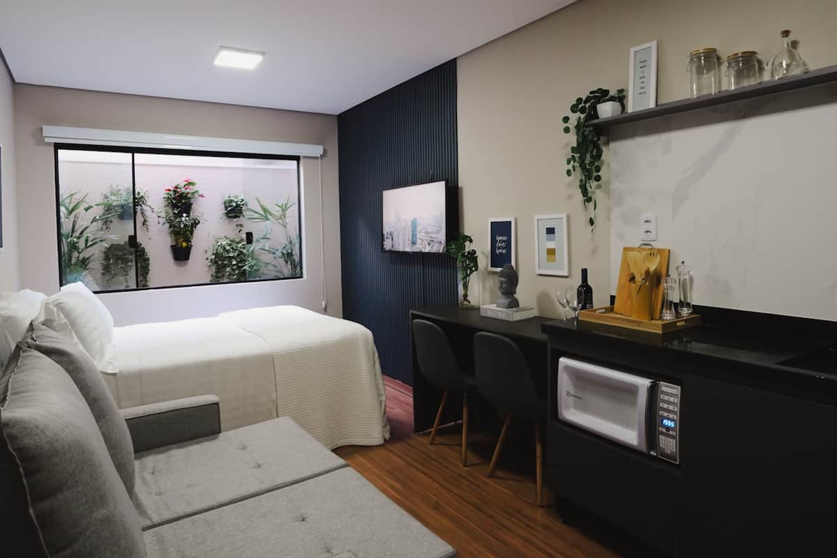 A compact hotel room with a bed, a desk, a chair, a small sofa, a window with plants, a TV, and a microwave.