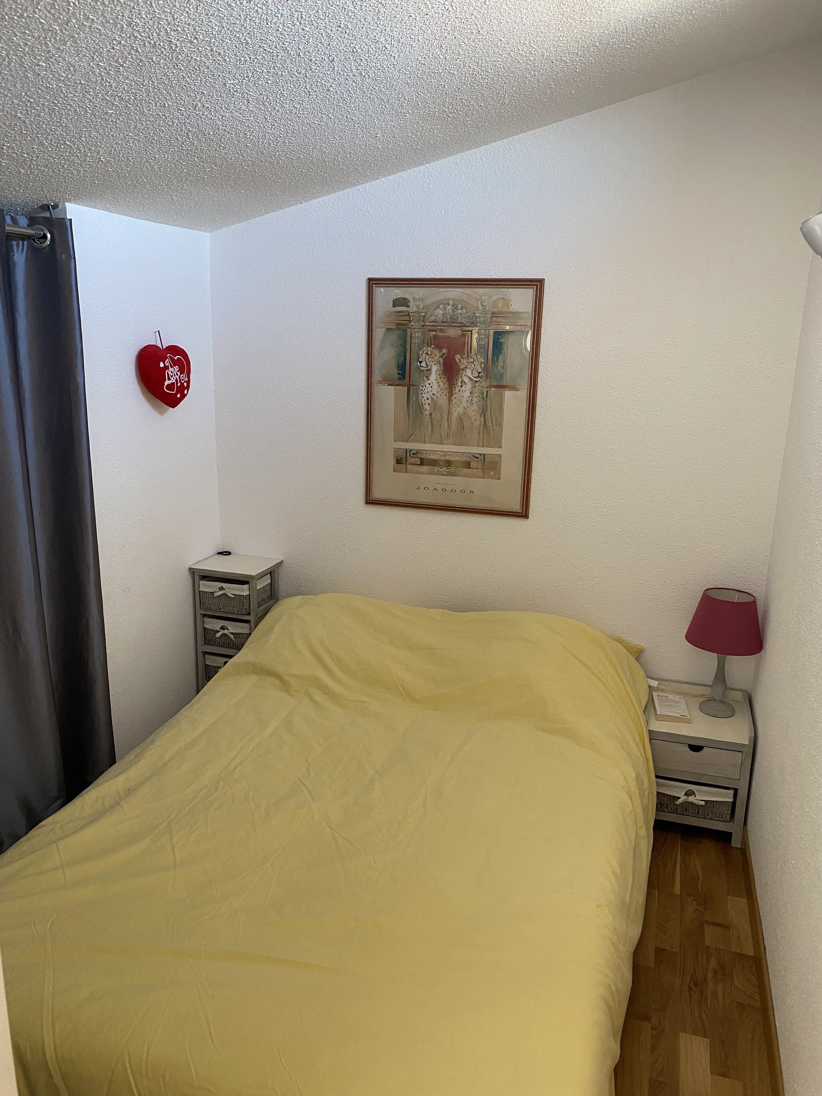 A bedroom with a bed, a nightstand, a lamp, a framed picture, a red heart decoration, and a small cabinet.