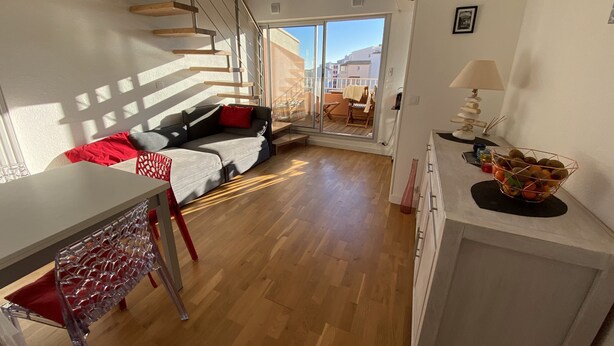 Een moderne woonkamer met een houten trap, een bank met rode kussens, en een eethoek met een tafel en stoelen.