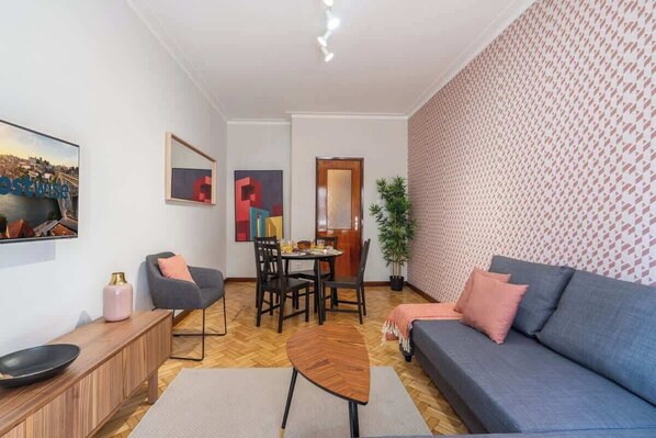 A modern living room with a grey sofa, a wooden coffee table, a grey armchair, and a dining table with chairs. There is a potted plant and a framed picture on the wall.