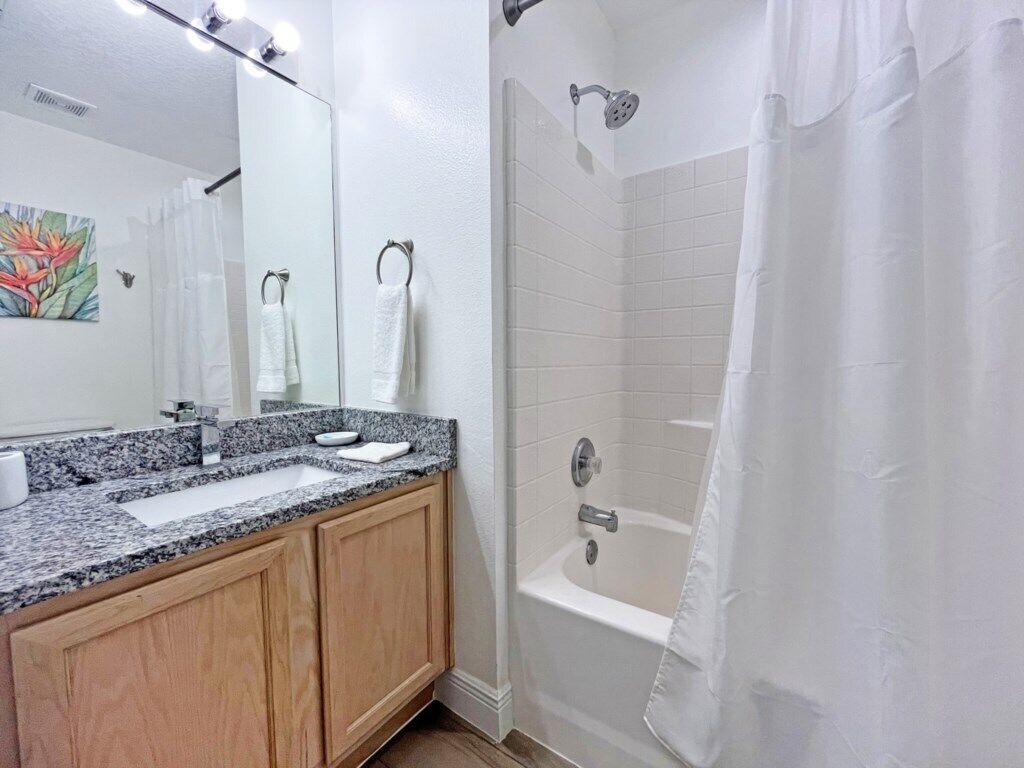 A bathroom with a granite countertop sink, wooden cabinets, a white bathtub, and a shower curtain.