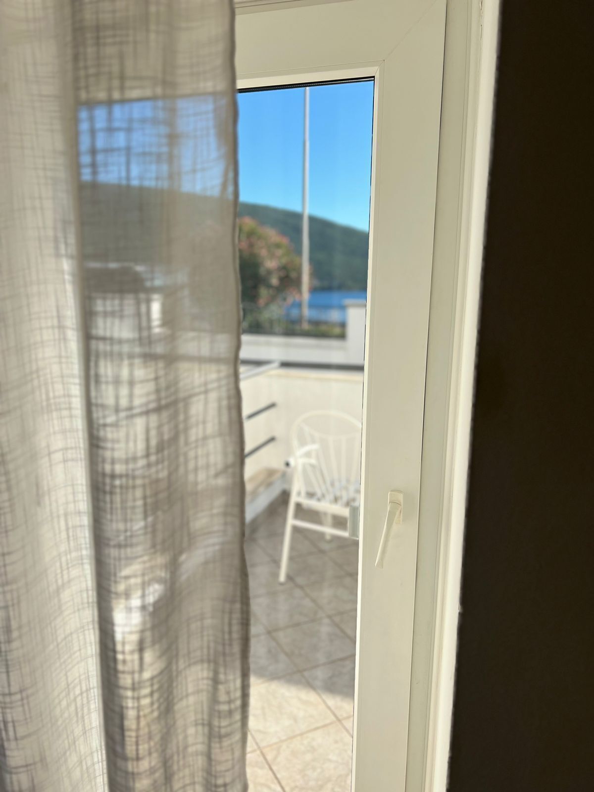 Vista a través de una puerta con cortina translúcida, que da a un espacio exterior con una silla blanca y una mesa, bajo un cielo azul despejado.